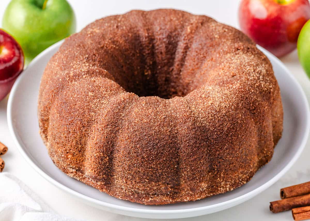 Apple cider donut cake on a white cake plate with fresh apples and cinnamon sticks in the background.