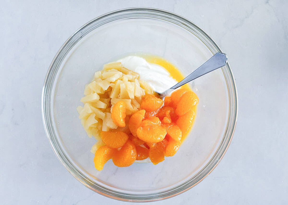 Glass bowl filled with chopped pineapple, mandarin oranges, and sour cream ready to be mixed for 5 cup salad.
