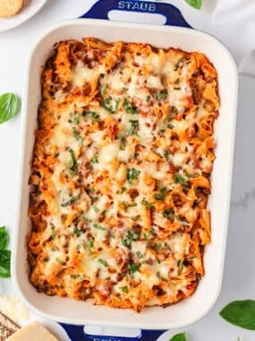 Baked lasagna casserole in a white Staub baking dish topped with melted cheese and fresh basil.