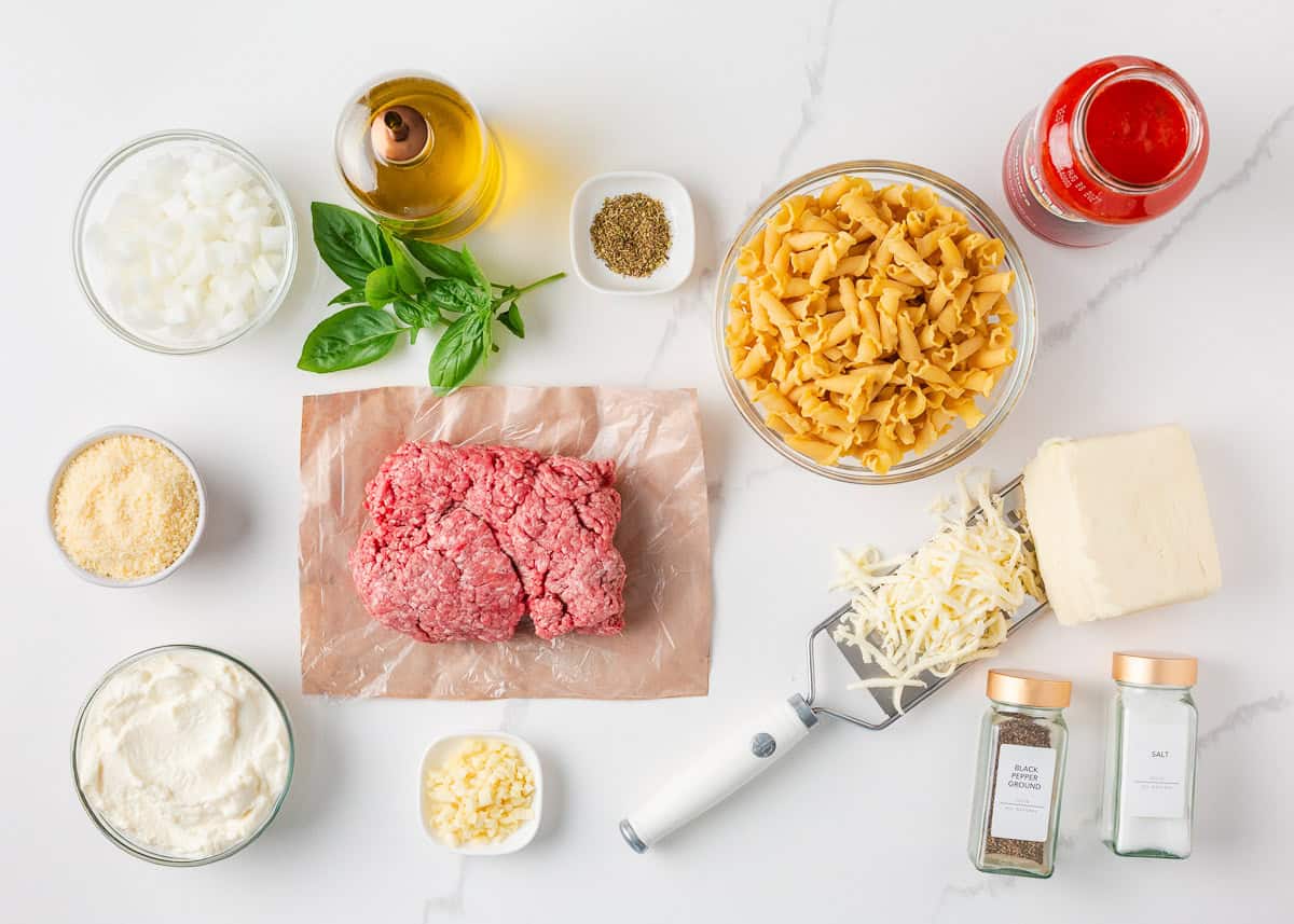 Ingredients for easy lasagna casserole arranged on a white surface, including ground beef, pasta, ricotta, marinara sauce, cheese, onion, garlic, and seasonings.