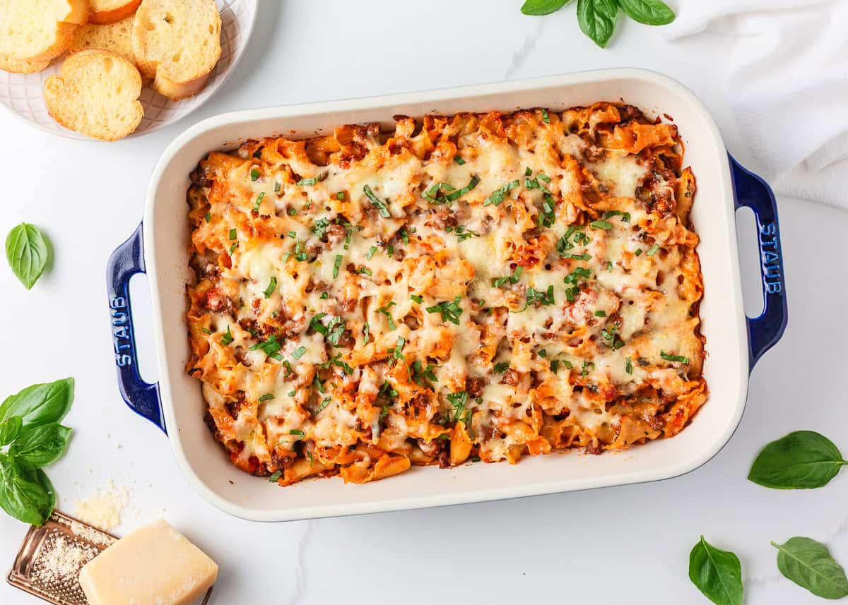 This lasagna casserole has all the classic flavors of traditional lasagna but in an easy-to-make casserole. This family-friendly baked pasta dish is perfect for weeknight dinners! Overhead view of an easy lasagna casserole in a white baking dish with golden melted cheese, surrounded by fresh basil and Parmesan.