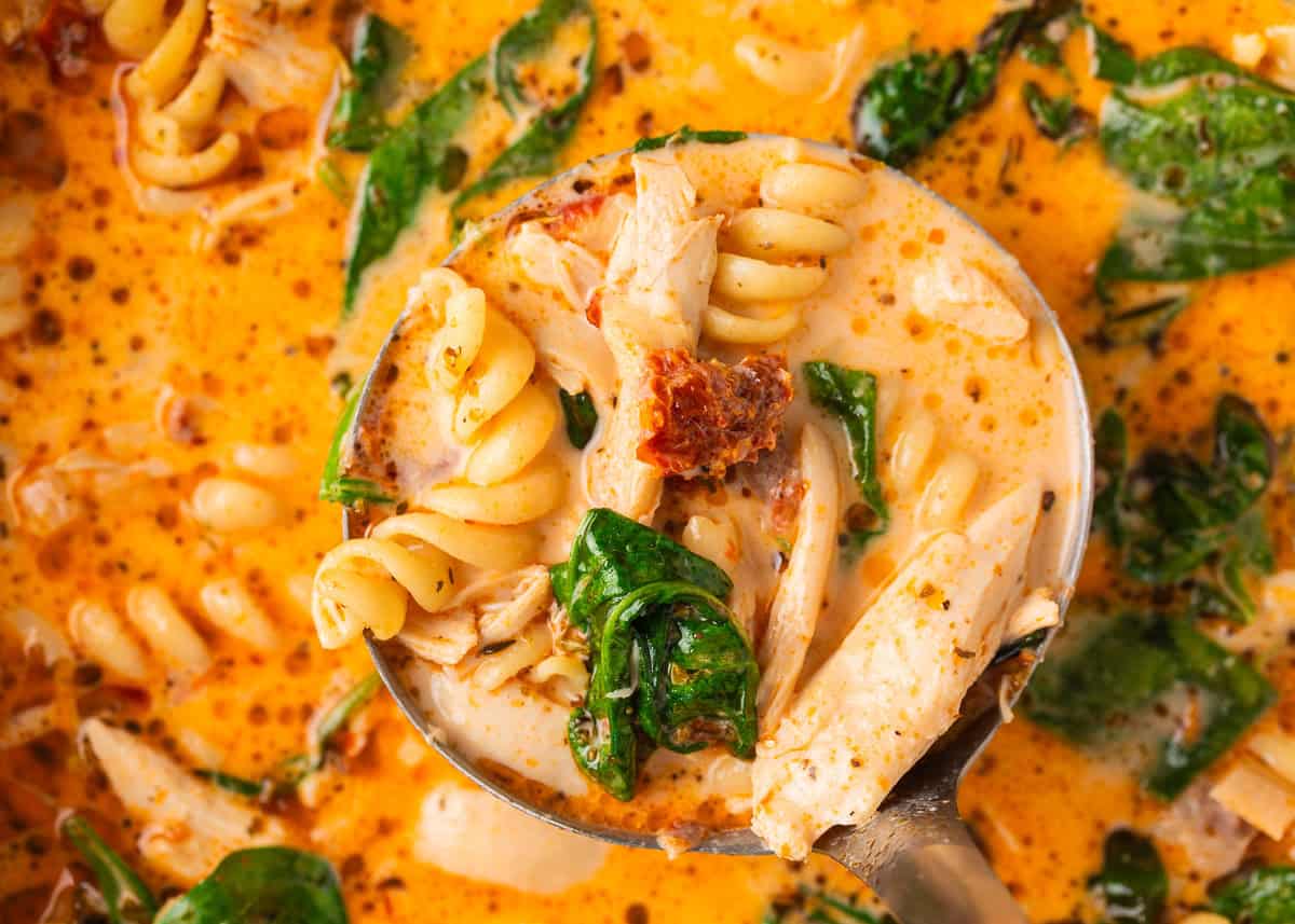 Marry me chicken soup is a cozy spin on the viral marry me chicken recipe made with tender chicken, pasta, sun-dried tomatoes and spinach in a Parmesan cream broth. Close-up of a ladle lifting creamy Marry Me Chicken Soup with rotini pasta, shredded chicken, spinach, and sun-dried tomatoes.