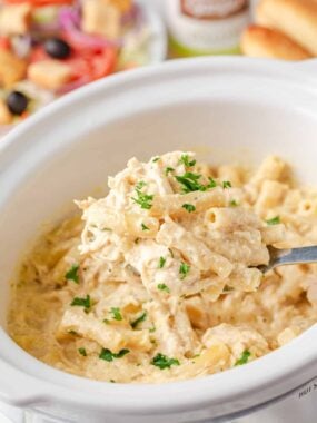 Spoonful of creamy crockpot Olive Garden chicken pasta held over slow cooker with salad in background.