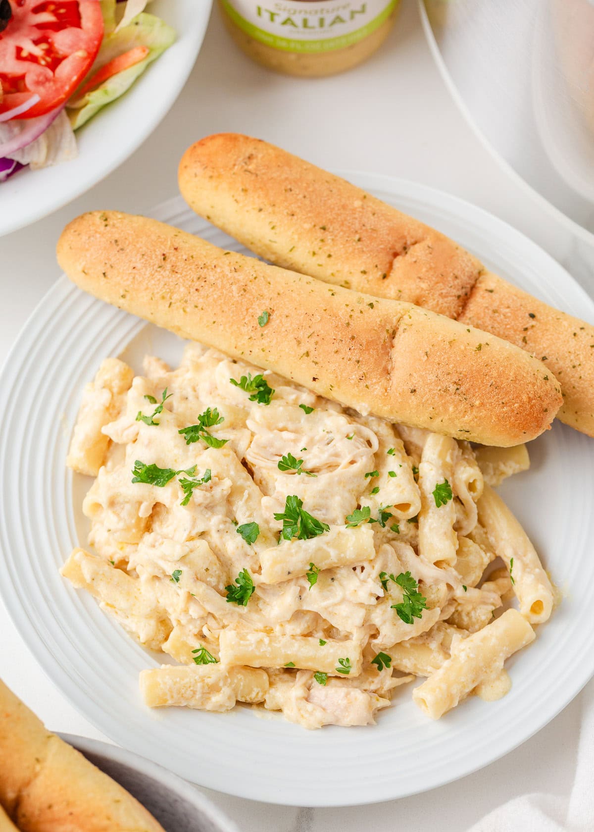 Olive Garden chicken pasta combines tender chicken slow-cooked in a rich, creamy, zesty sauce served over your favorite pasta, for the perfect weeknight meal at home! Serving of Olive Garden chicken pasta on a white plate with breadsticks and parsley.