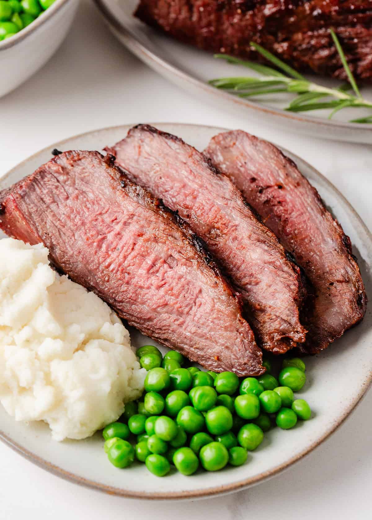 3 slices of tri tip on a plate with mashed potatoes and peas.