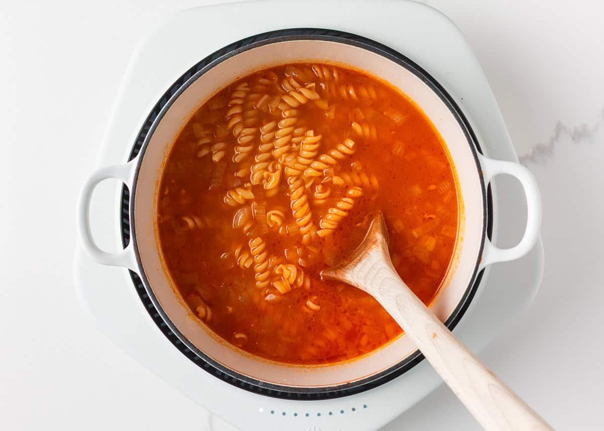Marry me chicken soup is a cozy spin on the viral marry me chicken recipe made with tender chicken, pasta, sun-dried tomatoes and spinach in a Parmesan cream broth. Step showing pasta simmering in tomato broth in a white pot on the stove.
