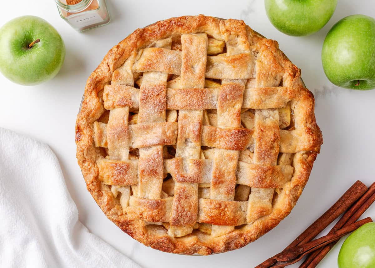 Baked apple pie on the counter with fresh green apples and cinnamon sticks.