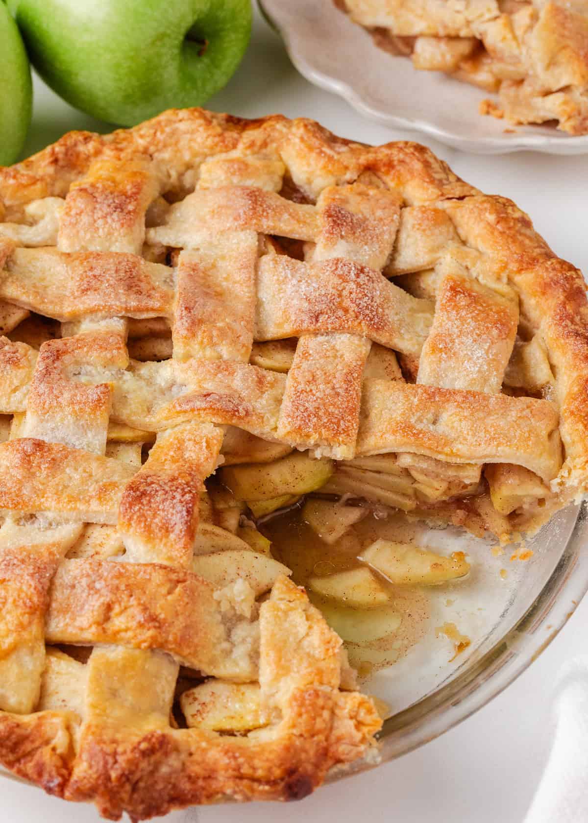 A baked apple pie with a lattice crust, slightly sliced to reveal the gooey cinnamon-apple filling, with green apples and a plate of pie in the background.