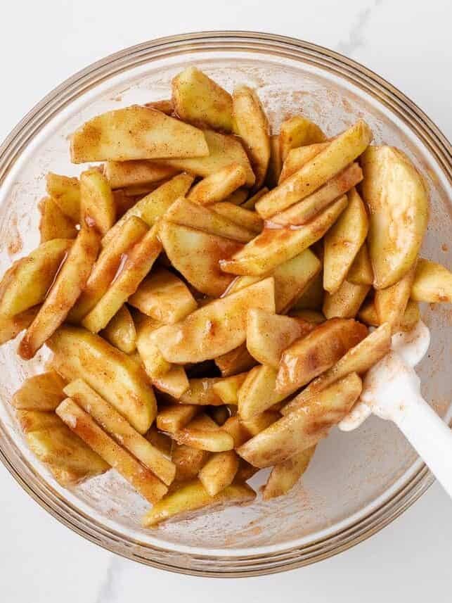 Overhead view of sliced green apples coated in cinnamon-sugar mixture in a glass bowl with a white spatula.