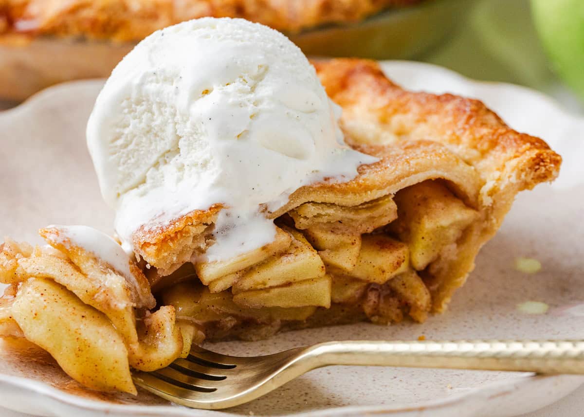 With a flaky pie crust and sweet, tart apples in every bite, this apple pie recipe is an easy dessert for Thanksgiving, Christmas, or anytime your family wants a taste of homemade. Close-up of a slice of apple pie with vanilla ice cream on top, fork cutting into the soft cinnamon-spiced apple filling.