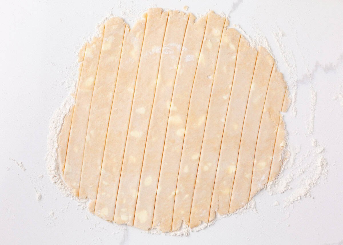 Pie dough rolled out and sliced into even strips on a floured surface, ready to be used for a lattice crust.