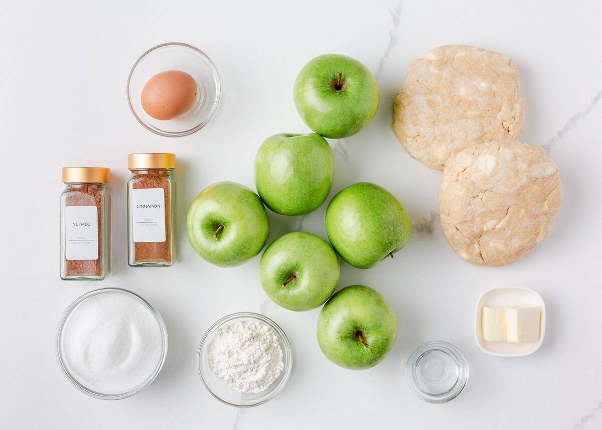 Ingredients for homemade apple pie including green apples, pie dough, egg, flour, sugar, butter, cinnamon, nutmeg, and water on a white background.