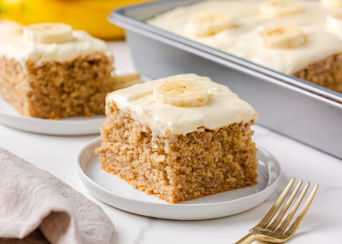 Easy banana cake with cream cheese frosting served in generous squares, topped with fresh banana and surrounded by ripe bananas.