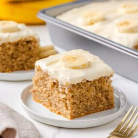 Thick slice of the best banana cake with creamy frosting and fresh banana garnish, with the full cake in the background.