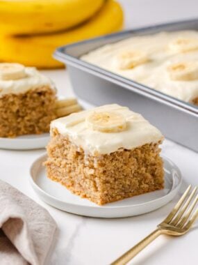 Thick slice of the best banana cake with creamy frosting and fresh banana garnish, with the full cake in the background.