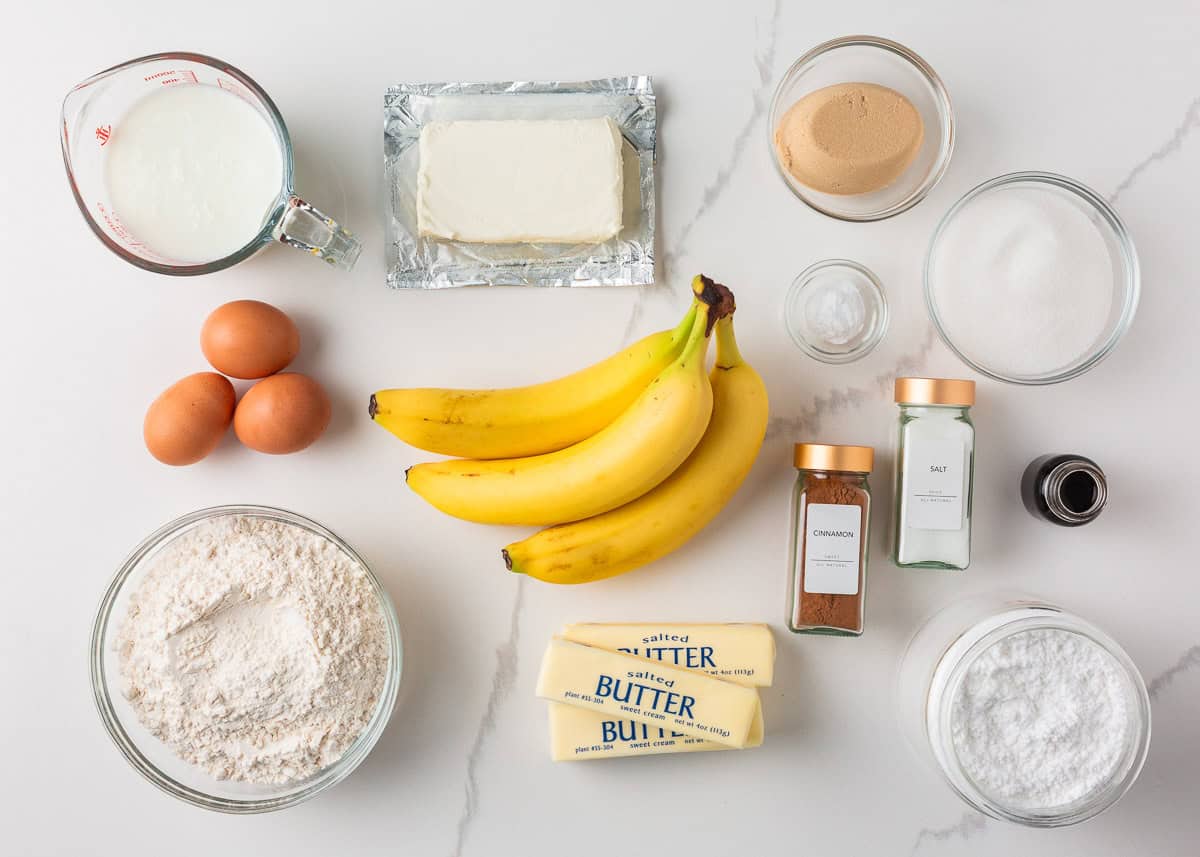 Ingredients for easy banana cake recipe including ripe bananas, flour, butter, cinnamon, eggs, cream cheese, and sugar arranged on a white counter.