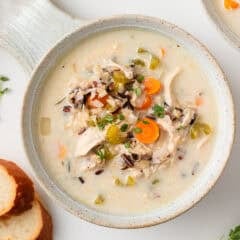 Overhead shot of two ceramic bowls filled with creamy chicken and wild rice soup, paired with fresh herbs and sliced baguette.