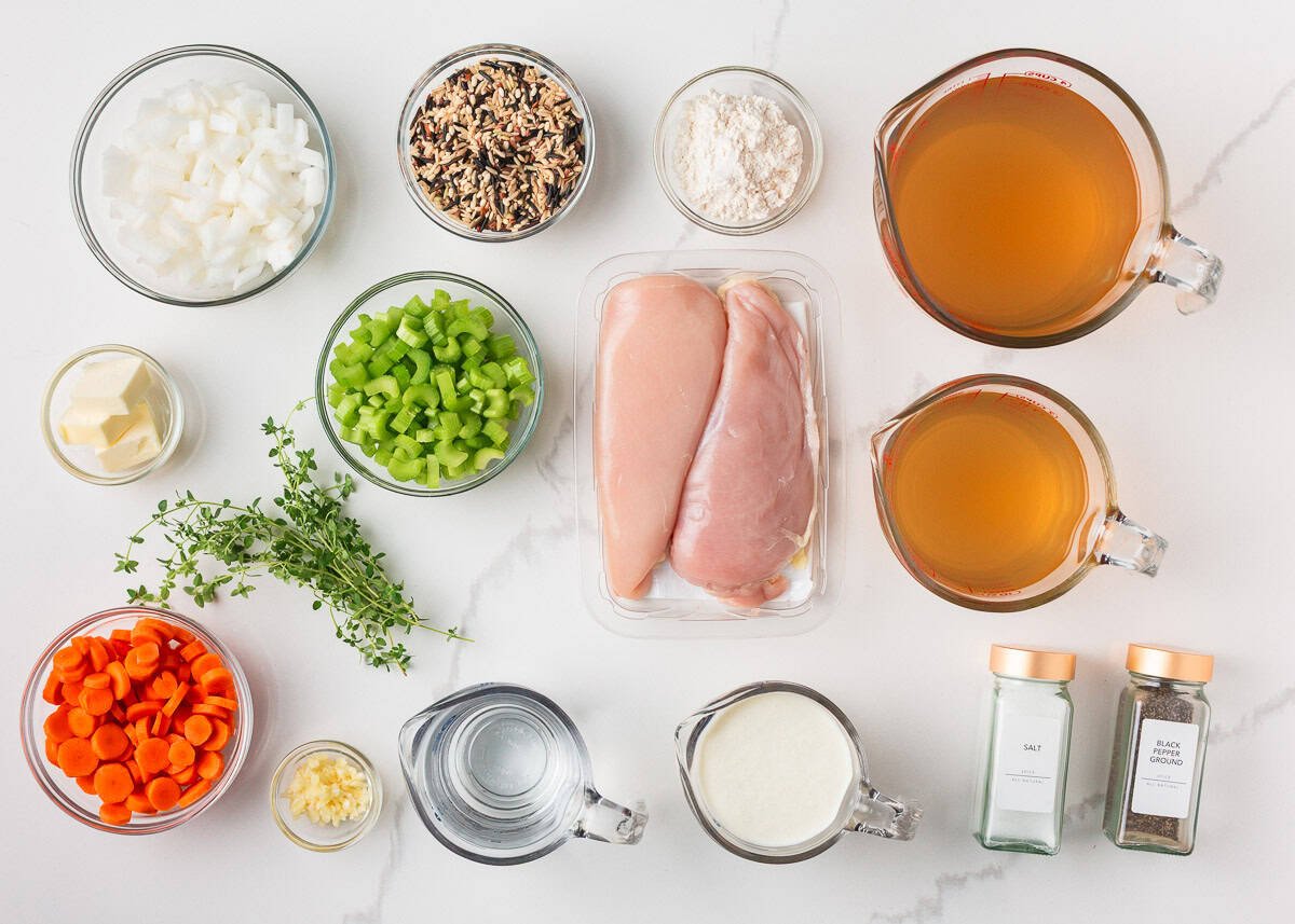 Flat lay of ingredients for creamy chicken and wild rice soup, including chicken breasts, carrots, celery, onions, garlic, wild rice, broth, cream, butter, flour, and fresh thyme.