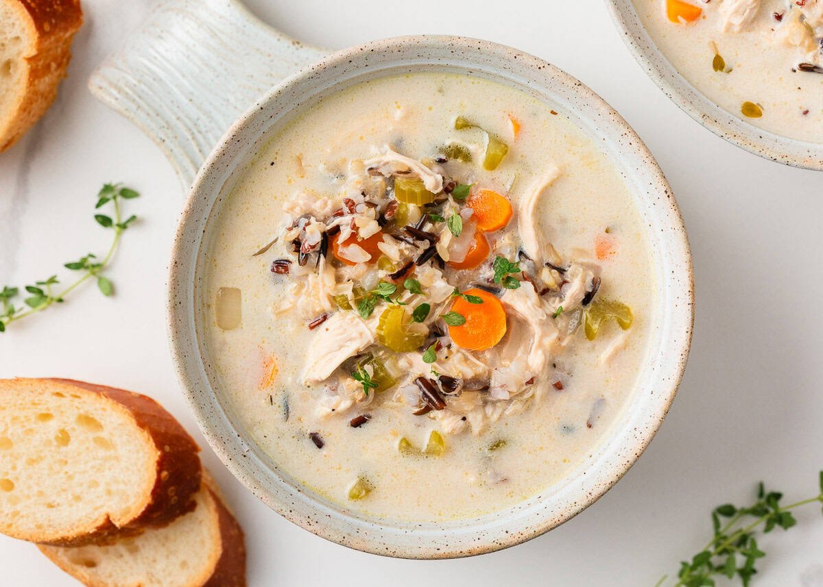 Overhead shot of two ceramic bowls filled with creamy chicken and wild rice soup, paired with fresh herbs and sliced baguette.