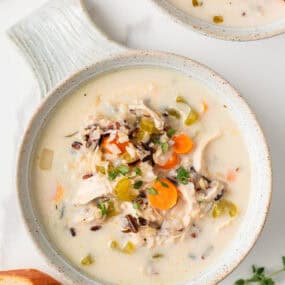 Top view of a bowl of creamy chicken and wild rice soup garnished with fresh thyme, surrounded by slices of crusty bread.