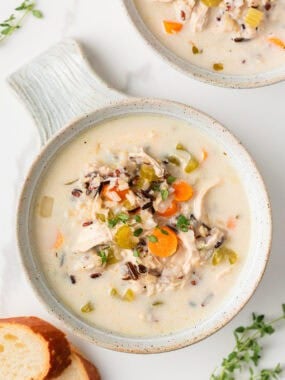 Top view of a bowl of creamy chicken and wild rice soup garnished with fresh thyme, surrounded by slices of crusty bread.