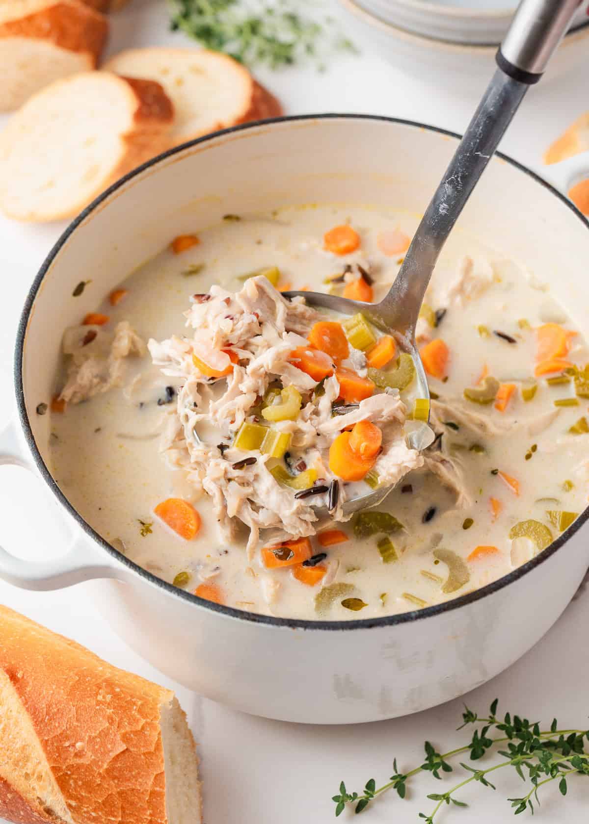 Overhead view of a Dutch oven filled with creamy chicken and wild rice soup, with a ladle lifting a hearty scoop and sliced baguette nearby.