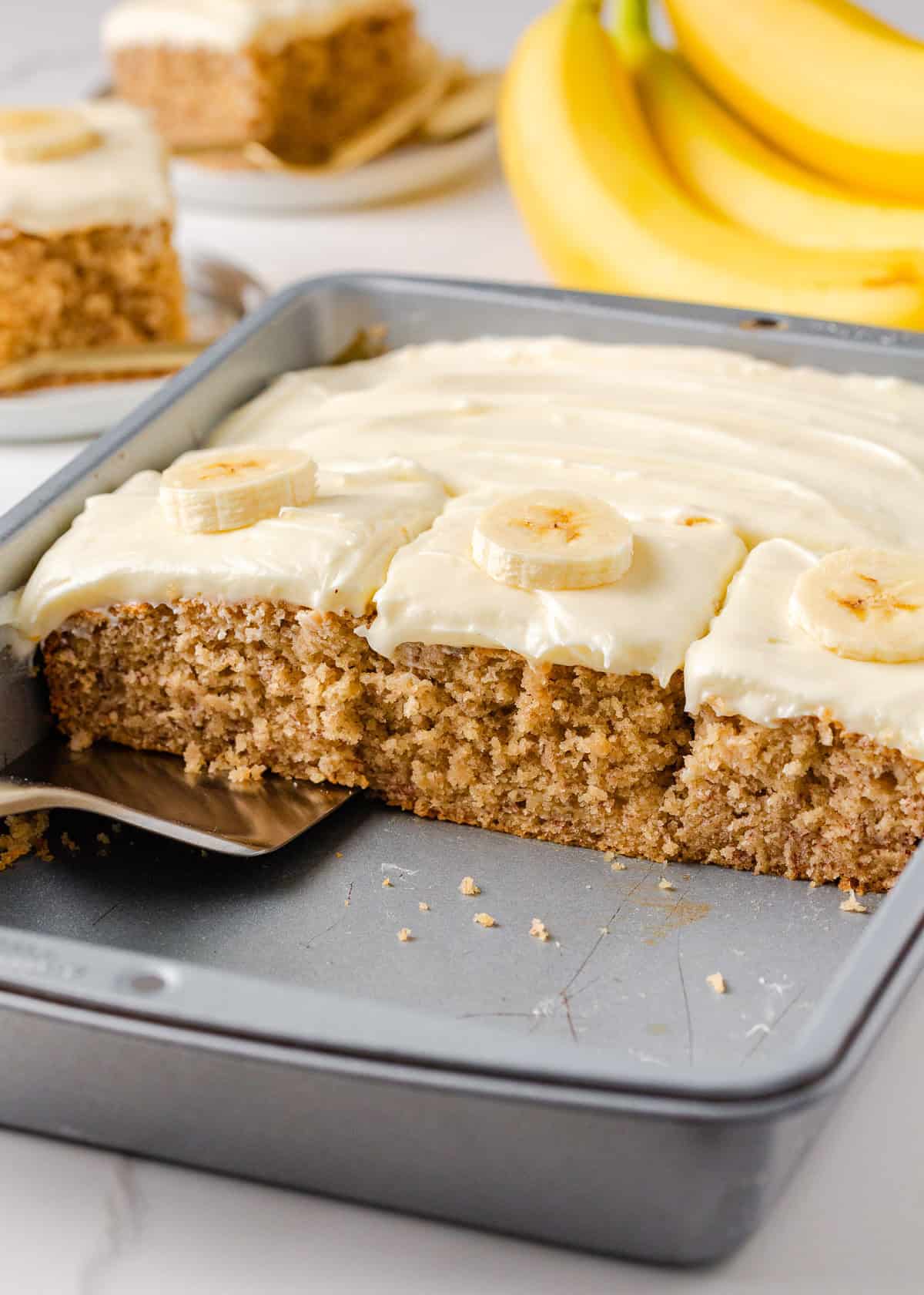 Banana cake with cream cheese frosting freshly sliced in the pan, revealing its soft, tender crumb and creamy topping.