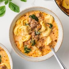 Bowl of Italian sausage tortellini soup topped with Parmesan cheese and surrounded by fresh basil leaves.