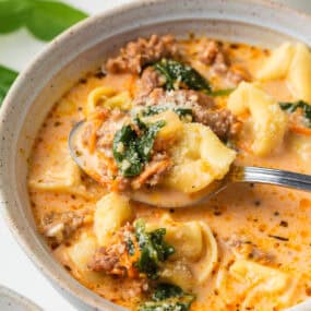 Closeup of a spoonful of Italian sausage tortellini soup lifted from a bowl, showing cheesy tortellini, browned sausage, wilted spinach, and creamy tomato broth.