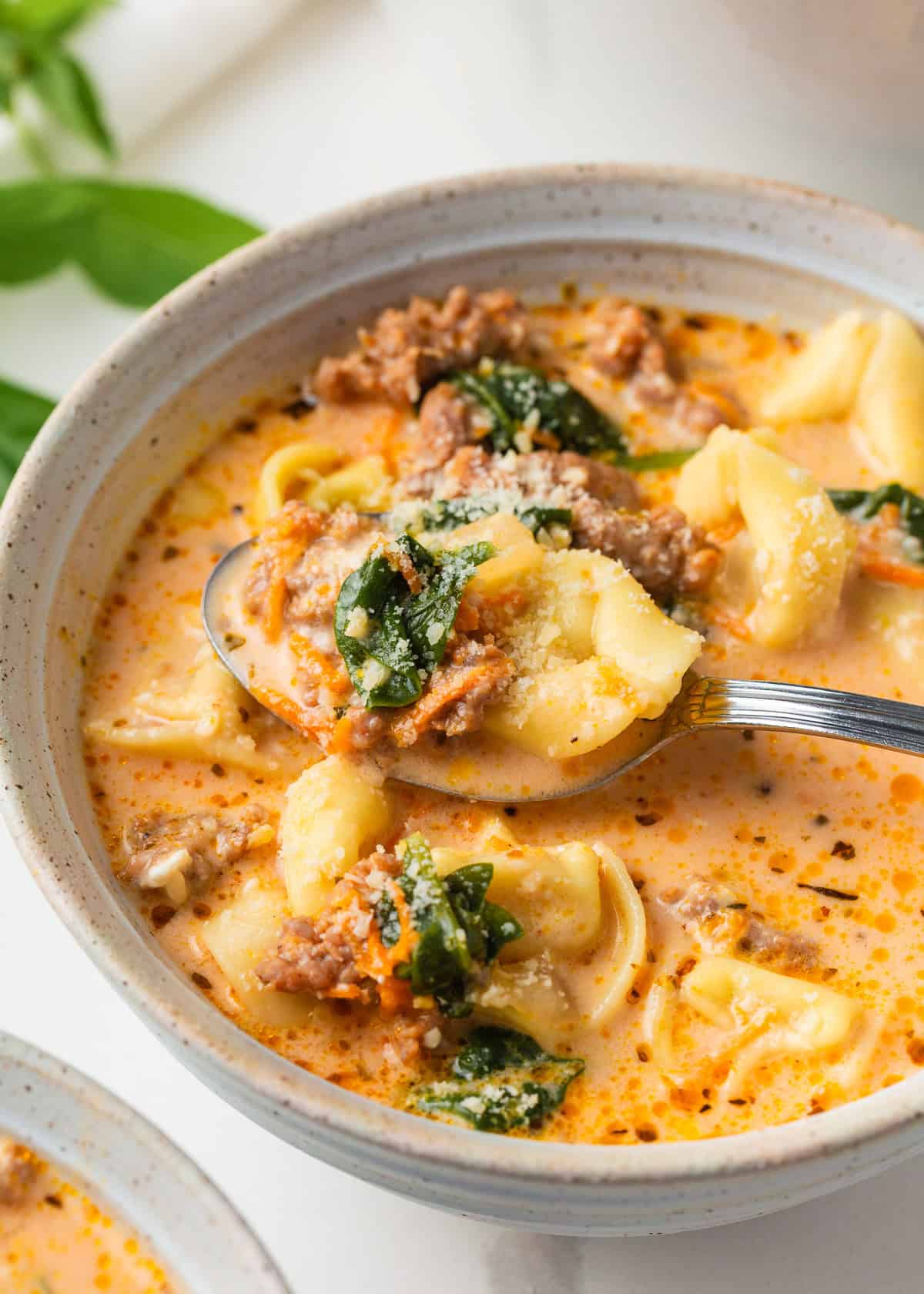 Closeup of a spoonful of Italian sausage tortellini soup lifted from a bowl, showing cheesy tortellini, browned sausage, wilted spinach, and creamy tomato broth.