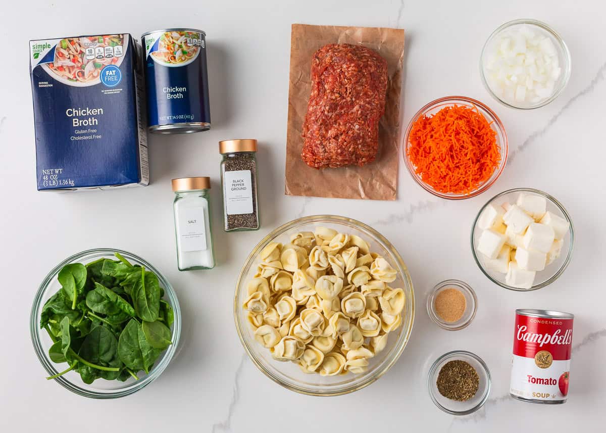 Packed with savory sausage, cheesy tortellini, and spinach in a creamy tomato broth, this Italian sausage tortellini soup is the perfect one-pot meal for a cozy family dinner! Ingredients for Italian sausage tortellini soup arranged on a white counter, including tortellini, ground sausage, spinach, carrots, broth, cream cheese, tomato soup, onion, and seasonings.