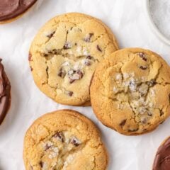 Jacque Torres chocolate chip cookies topped with flaky sea salt and some dipped in melted chocolate, arranged on parchment paper.