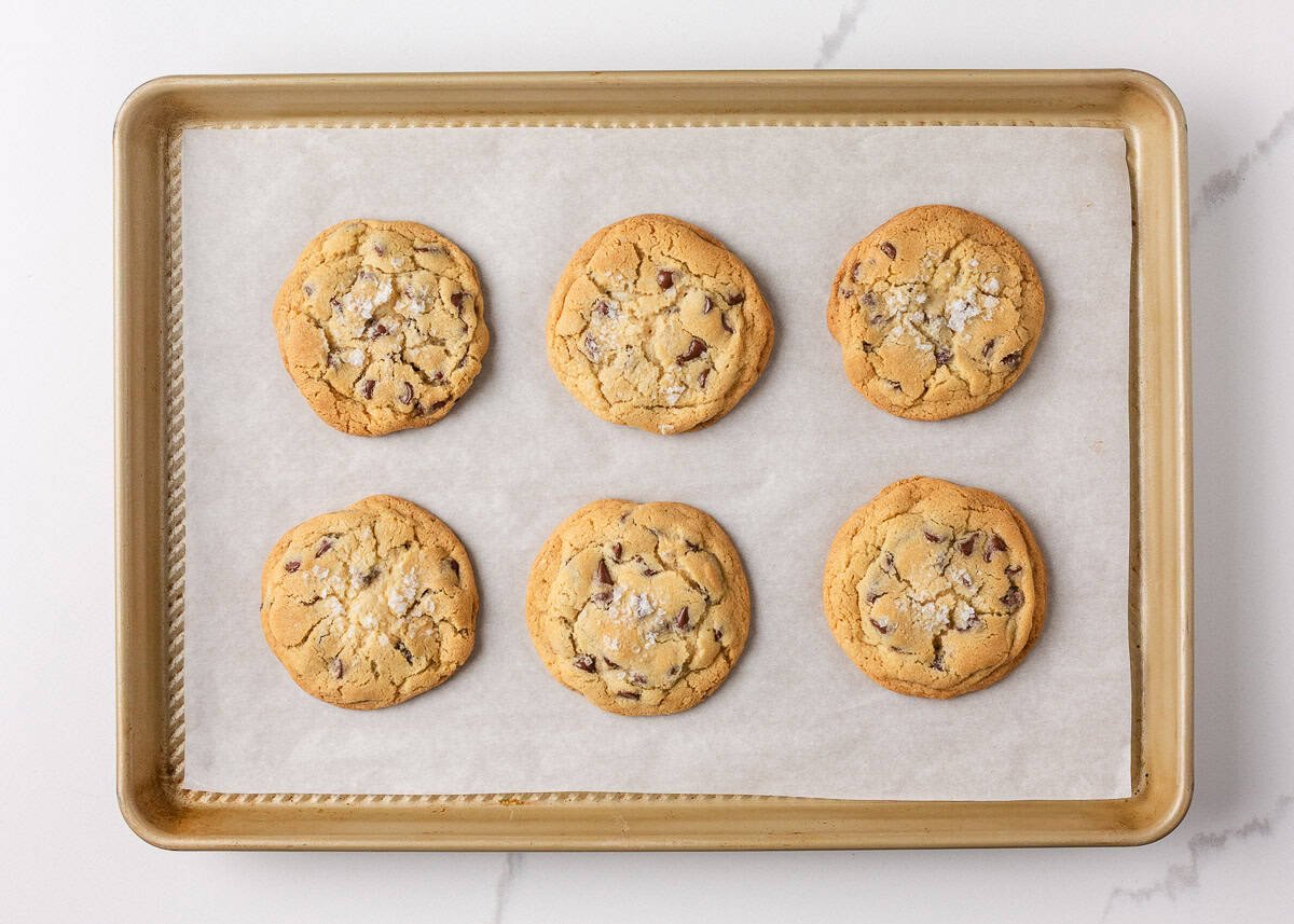 Six Jacque Torres chocolate chip cookies fresh out of the oven on a parchment-lined baking sheet, golden brown with sea salt sprinkled on top.