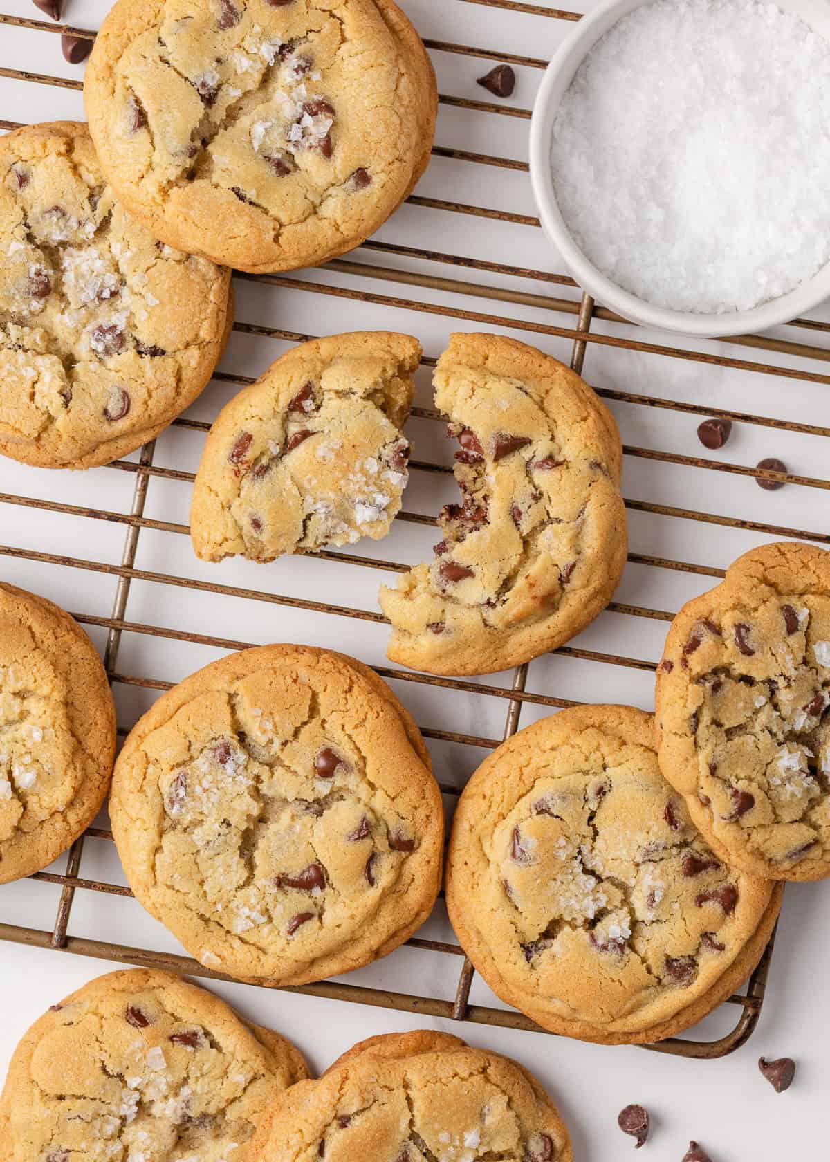Freshly baked Jacque Torres chocolate chip cookies cooling on a wire rack, sprinkled with sea salt and surrounded by chocolate chips.
