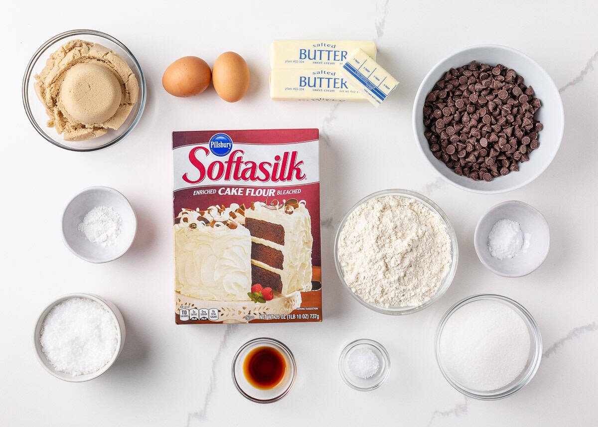 Flat lay of ingredients for Jacque Torres chocolate chip cookies, including cake flour, brown sugar, butter, eggs, small chocolate discs, and vanilla.
