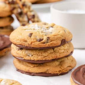 Stack of Jacque Torres chocolate chip cookies with melted chocolate bottoms and sea salt topping, with blurred cookies in the background.