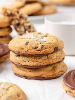 Stack of Jacque Torres chocolate chip cookies with melted chocolate bottoms and sea salt topping, with blurred cookies in the background.