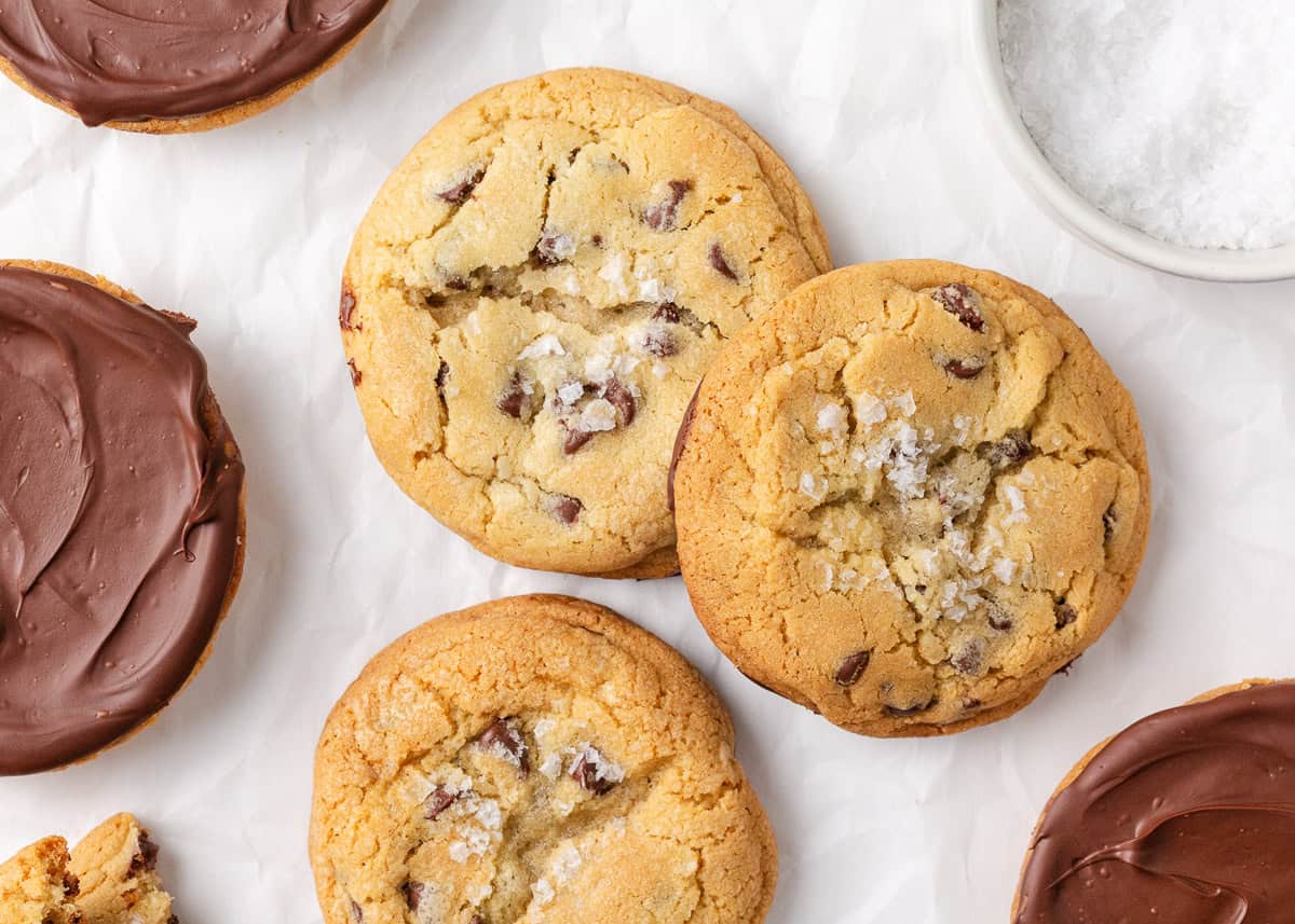 Jacque Torres chocolate chip cookies topped with flaky sea salt and some dipped in melted chocolate, arranged on parchment paper.