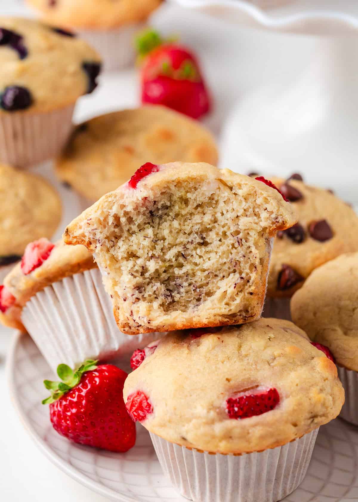 A close-up of a pancake muffin with a bite taken out, showing the moist, tender inside. Other muffins with strawberries and chocolate chips are arranged on the plate.