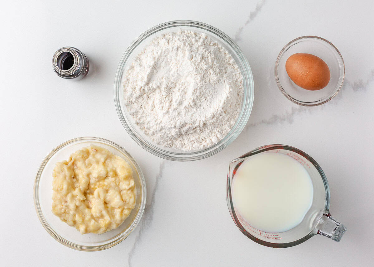 Ingredients for pancake muffins arranged on a counter, including mashed banana, flour, milk in a measuring cup, an egg, and a bottle of vanilla extract.