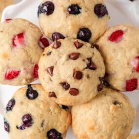 Overhead view of assorted pancake muffins topped with chocolate chips, blueberries, and strawberries arranged in a circle on a white platter.