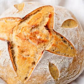 Overhead view of a round loaf of same day sourdough bread with a star and leaf scoring pattern, resting on a white cloth.