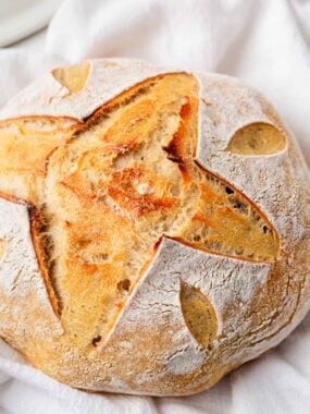 Overhead view of a round loaf of same day sourdough bread with a star and leaf scoring pattern, resting on a white cloth.