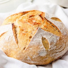 Golden brown round loaf of same day sourdough bread with decorative scoring, placed on a soft white kitchen towel.