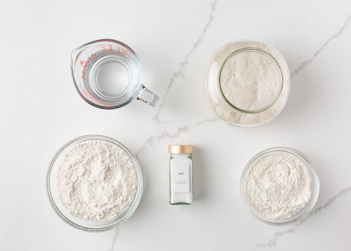 Flat lay of same day sourdough bread ingredients including water, sourdough starter, flour, and salt arranged on a white marble surface.