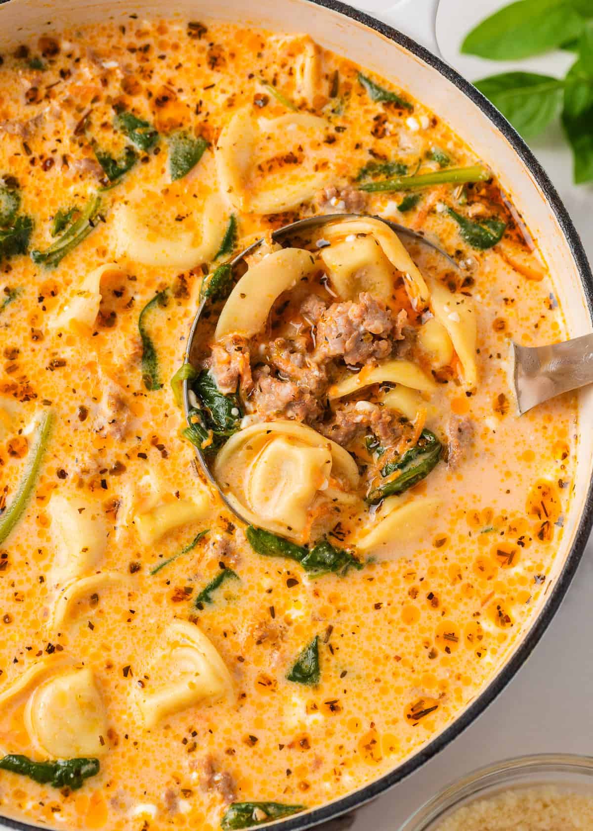 Packed with savory sausage, cheesy tortellini, and spinach in a creamy tomato broth, this Italian sausage tortellini soup is the perfect one-pot meal for a cozy family dinner! Closeup of Italian sausage tortellini soup in a white pot with a ladle scooping a hearty serving of tortellini, sausage, and spinach in a creamy tomato broth.