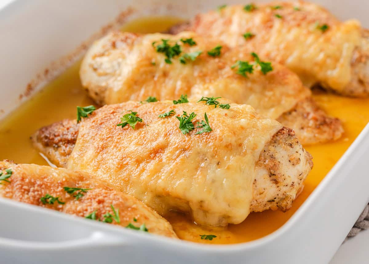 There is nothing better than Swiss chicken bake made with a handful of ingredients and a creamy, cheesy golden topping. Pair with rice and veggies, this easy dinner is served! Angled view of Swiss chicken breasts baked in a white dish garnished with parsley.