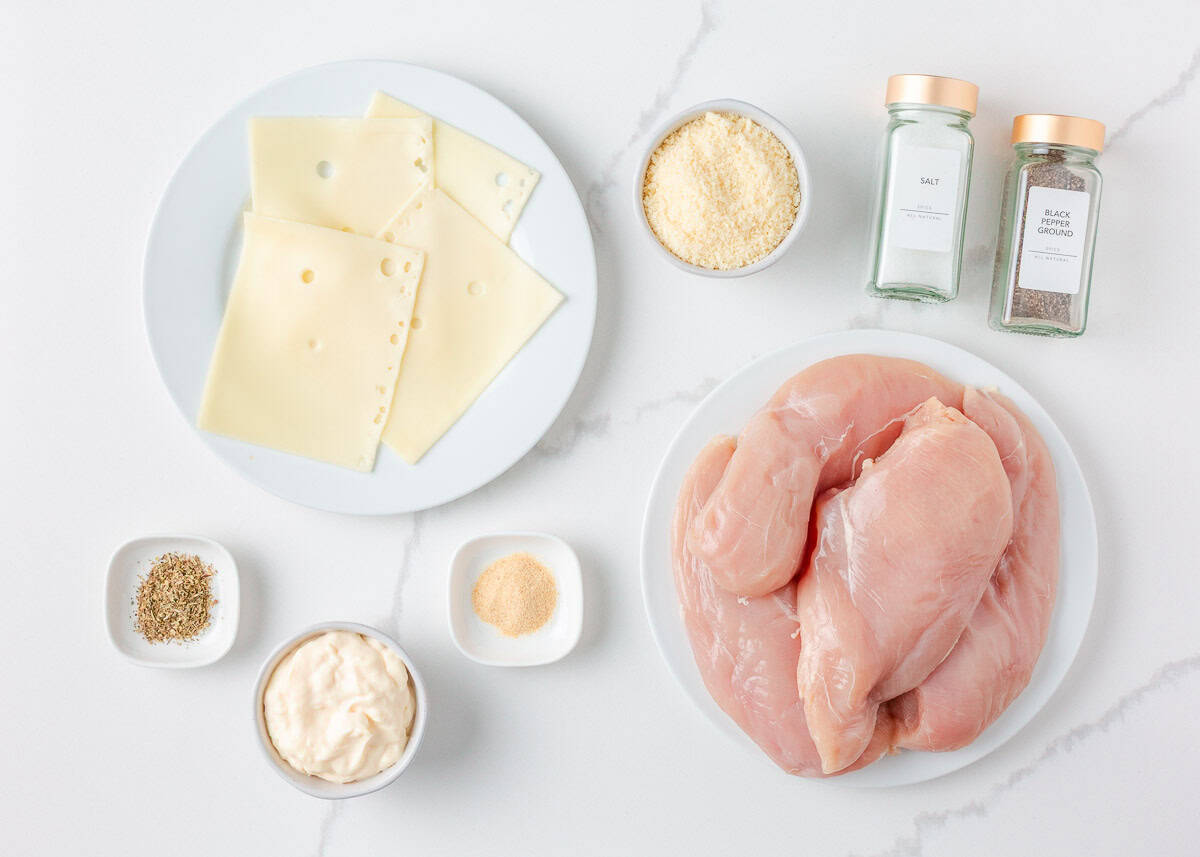 There is nothing better than Swiss chicken bake made with a handful of ingredients and a creamy, cheesy golden topping. Pair with rice and veggies, this easy dinner is served! Ingredients for Swiss chicken including raw chicken breasts, Swiss cheese slices, shredded parmesan, mayonnaise, spices, and seasonings arranged on a white surface.