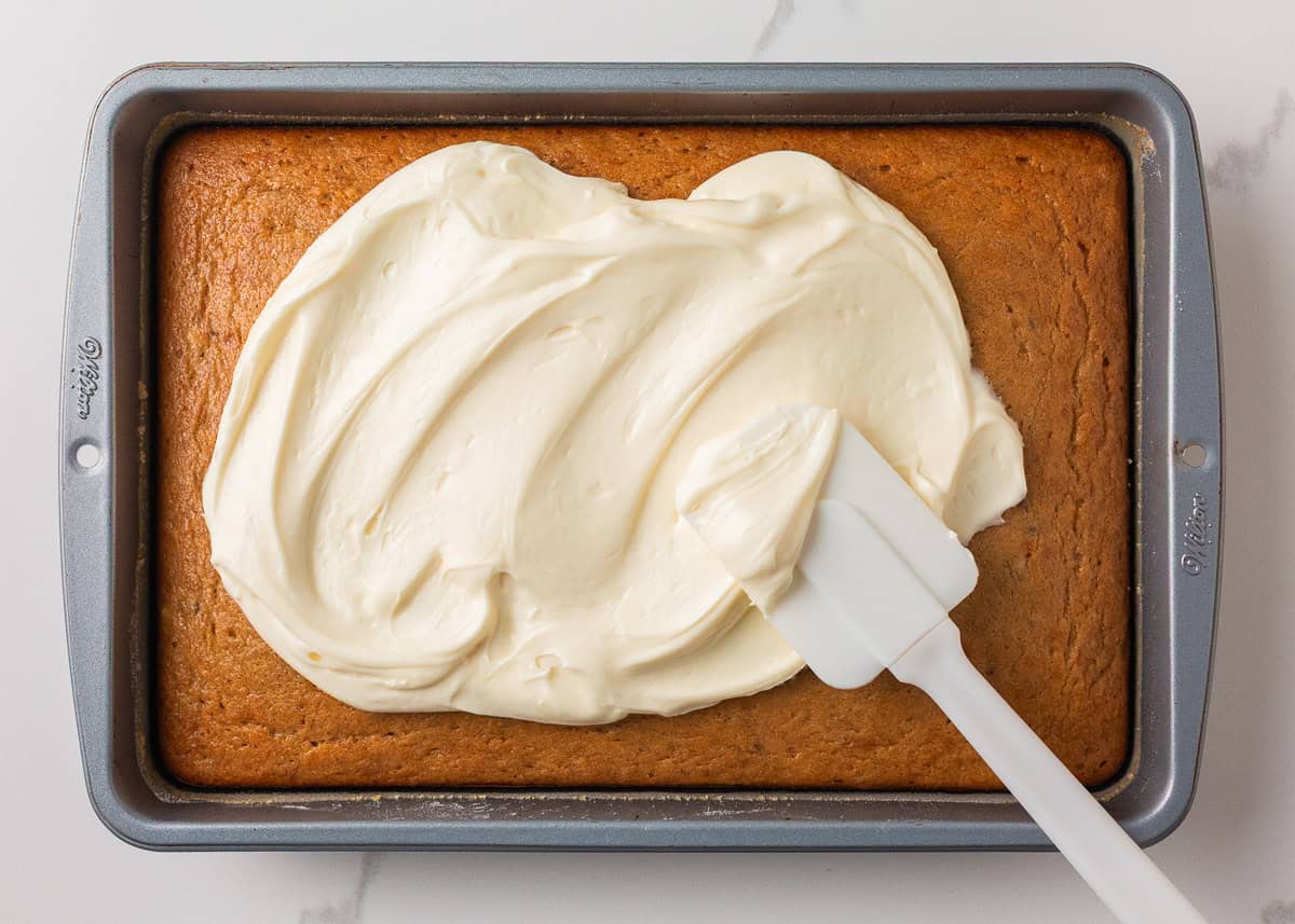 Cream cheese frosting being spread over the cooled banana cake with a spatula.