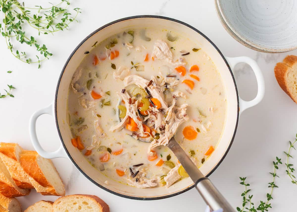Top-down view of a Dutch oven filled with creamy chicken and wild rice soup, with a ladle resting in the pot and herbs and bread scattered around.